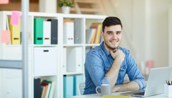 Happy young successful office worker in casualwear sitting by desk in front of camera and networking
