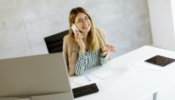 Pretty young woman using mobile phone while sitting by the desk in the office